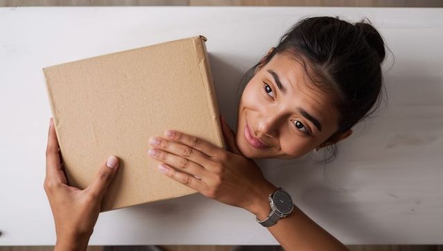 Receiving parcel through table opening by smiling Asian woman reaching for cardboard box