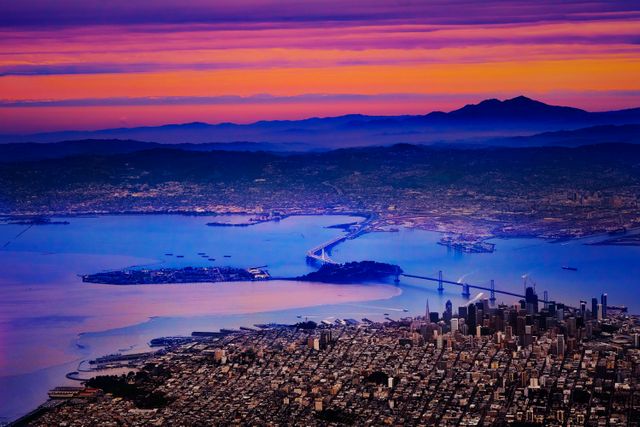 Vibrant aerial shot of San Francisco Bay and surrounding cityscape with Golden Gate Bridge in the background during sunset. Ideal for travel guides, tourism promotions, and cityscape photography collections. Highlights striking colors of the sky and picturesque waterfront details.
