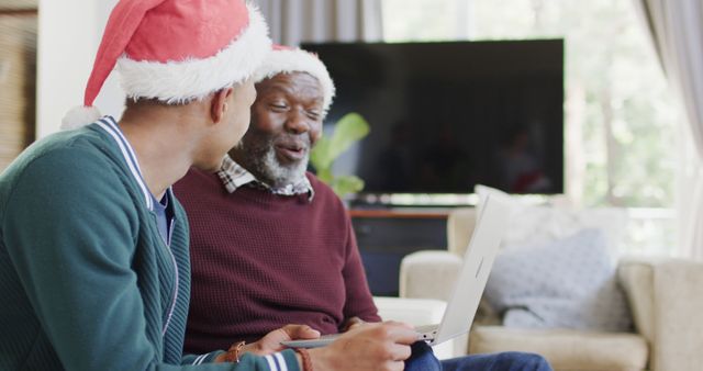 Father and son sharing joyful moment wearing Santa hats while using laptop. Great for holiday-themed promotions, family tech products, and festive greeting cards.