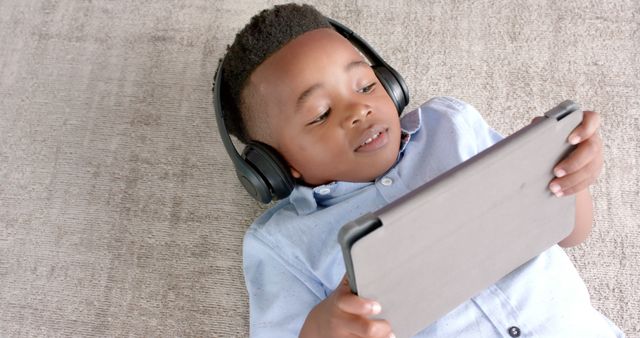 Young boy laying on carpet while using tablet and wearing headphones. Captures modern technology and childhood leisure time. Ideal for educational materials, tech advertisements, parenting blogs, or any content promoting children's technology engagement.