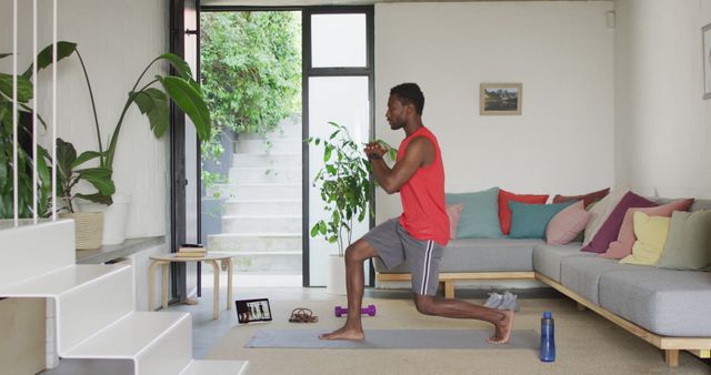 Man doing home workout in living room, performing lunge exercises on yoga mat. Casual fitness setup, including water bottle, tablet on floor, dumbbells. Use for fitness guides, home workout videos, active lifestyle promotions.