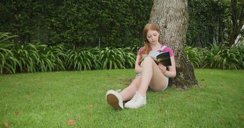 Teen Girl Reading Under Tree on Green Lawn with Backpack and White Sneakers