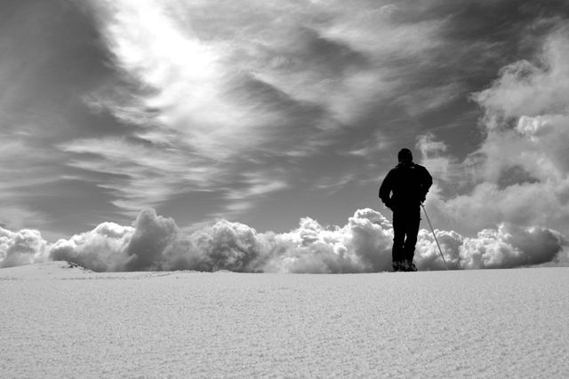 A solitary hiker is standing on snowy terrain, silhouetted against a dramatic, cloudy sky. This image evokes feelings of solitude and adventure, making it ideal for motivational posters, travel brochures, and winter sports advertisements.