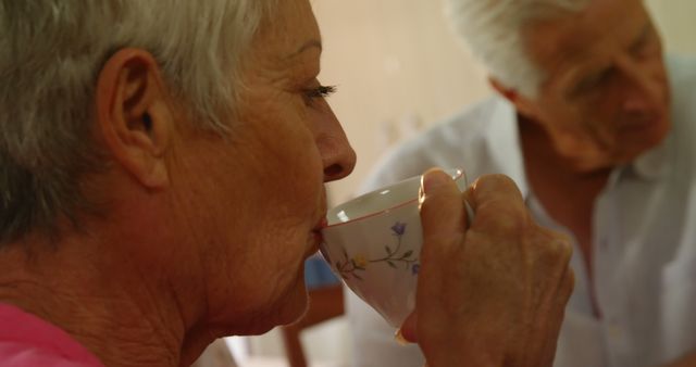 Senior woman enjoying tea while engaging with elderly man indoors. Featuring relaxed, cozy atmosphere. Perfect for illustrating themes of elderly lifestyle, retirement, daily routine of older adults, and health awareness.
