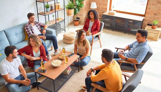 Friends chatting and laughing around coffee table in modern loft living room with snacks and beers