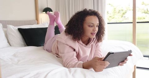African american woman lounging on bed in pink loungewear using tablet in bright bedroom