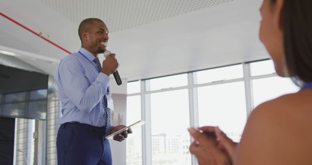 Businessman wearing formal attire, giving engaging presentation using a microphone in a modern office. Colleagues in audience applaud, creating an atmosphere of support and teamwork. Can be used for business communications, leadership training, teamwork presentations, and motivational speaking.