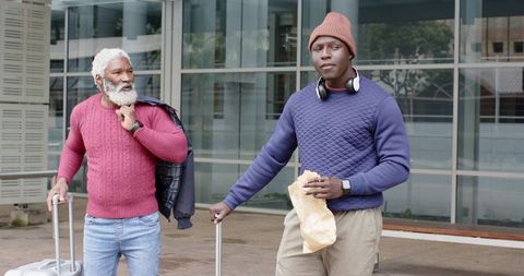 African American senior and adult men standing with luggage at modern urban glass entrance