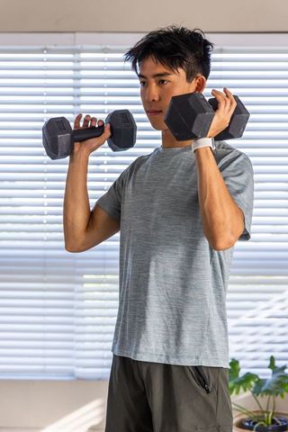 Asian man lifting dumbbells at home during daylight workout wearing fitness watch
