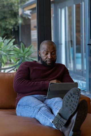 African american man reclining on leather sofa using tablet, relaxing in modern home