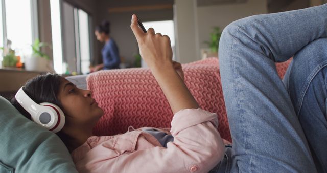 Young woman lying on a sofa at home and using her smartphone while listening to music with headphones. Suitable for illustrating concepts related to technology, relaxation, modern lifestyle, home comfort, and leisure activities.