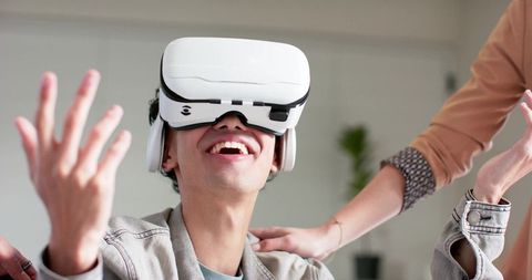 Young man wearing white VR headset smiling and reaching while friends guiding him at home