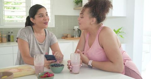 Friends chatting over pink smoothies at bright kitchen island, sharing healthy routine