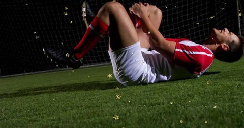 Soccer player clutching knee and lying on turf under floodlights near goal net