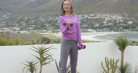 Mature woman exercising with purple dumbbells on coastal balcony overlooking bay