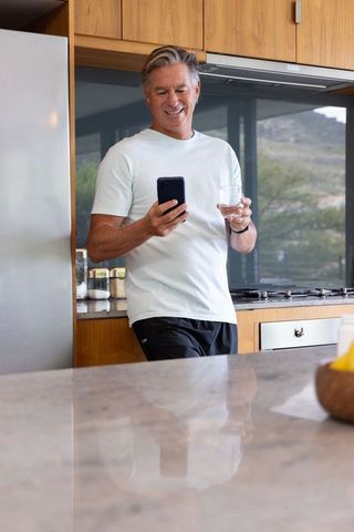 Mature man leaning at kitchen counter checking smartphone while holding glass of water