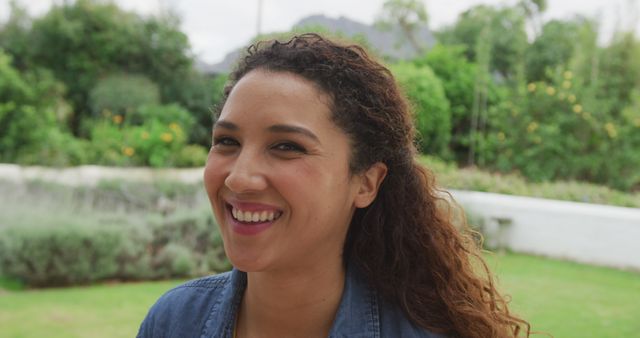 Woman with curly hair smiling genuinely outdoors. Ideal for use in content related to happiness, wellbeing, positive vibes, outdoor activities, and wellness promotions.