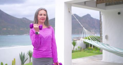 Mature woman lifting dumbbells on coastal patio wearing magenta top and smartwatch