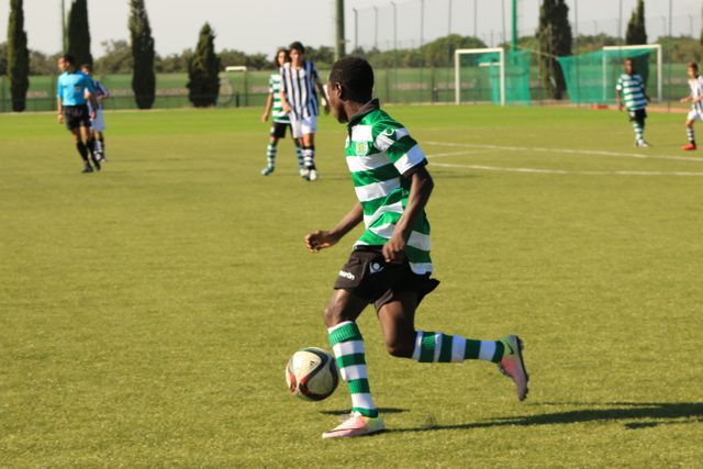 Youth soccer player in green and white uniform dribbling ball on grass field during a match. Other players in the background visible. Ideal for depicting youth sports, teamwork, and soccer-related content.