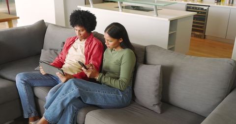 African American and Indian Couple Relaxing on Gray Sofa Using Tablet and Phone