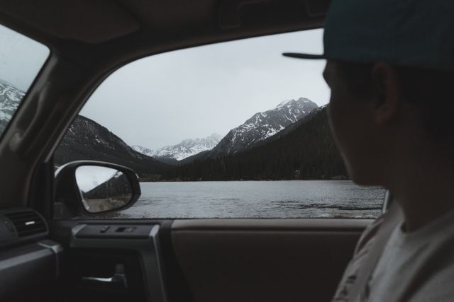 Person sitting inside a car, enjoying a view of a scenic mountain lake. Useful for content about travel, adventure, nature exploration, road trips, and serene outdoor landscapes.