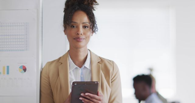 Young African American businesswoman standing confidently in office holding tablet. Professional work attire shows readiness and competence. Suitable for business, corporate culture, workplace productivity, or technology in business-related visuals. Great for websites, brochures, presentations, and advertising materials.