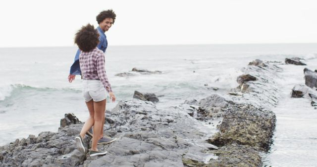 Enthusiastic young couple enjoying a carefree moment walking on a rocky seashore near the ocean. Ideal for themes related to love, romance, adventure, vacations, and outdoor activities. Great for blogs, travel websites, relationship advice content, or social media posts focusing on joyful, scenic moments and togetherness.
