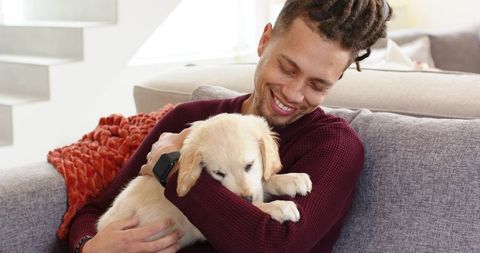 Smiling man cuddling golden retriever puppy on couch at home wearing burgundy sweater