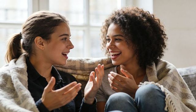 Teen girls laughing and talking on sofa wrapped in cozy blanket warm natural daylight