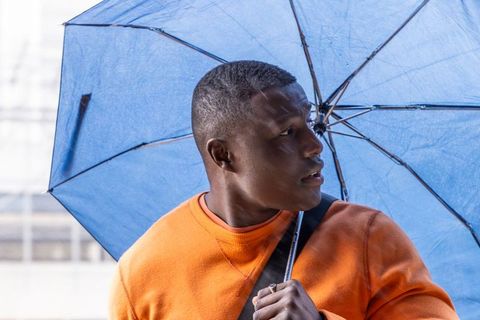 African american man holding blue umbrella wearing orange sweater looking to side