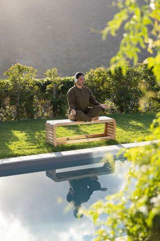 Asian woman meditating by pool on wooden bench wearing olive activewear and earbuds