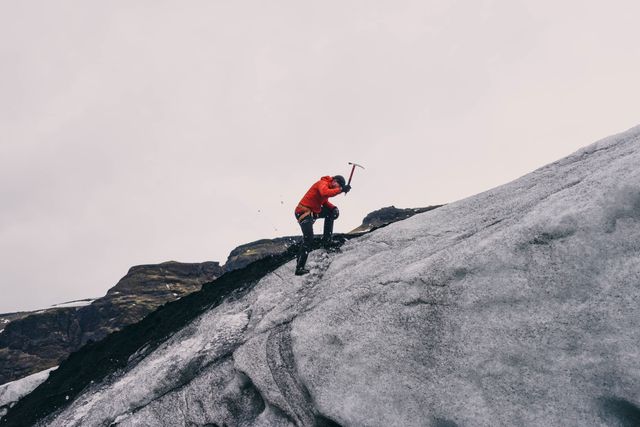 Man wearing red jacket and using an ice axe to climb a glacier in remote Icelandic location. Ideal for travel blogs, adventure magazine covers, extreme sports promotions, and outdoor activity advertisements.