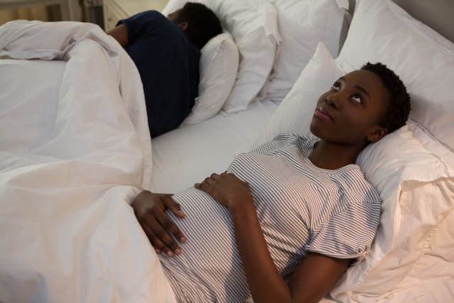 Pregnant woman lying in bed at home, looking thoughtful. Ideal for content related to pregnancy, maternity, relaxation, and domestic life. Can be used in articles, blogs, and advertisements focusing on expecting mothers, mental health during pregnancy, and home life.