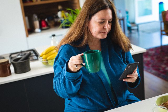 Caucasian mature woman using smart phone while sitting with coffee cup in kitchen at home - Download Free Stock Photos Pikwizard.com