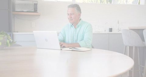 Mature man working remotely at bright modern kitchen table with laptop and notebook