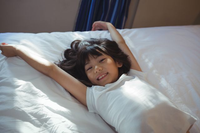 Young girl lying on bed with arms stretched out, smiling happily. Ideal for use in family, home, and lifestyle contexts, promoting comfort, relaxation, and childhood joy. Perfect for advertisements, blogs, and articles related to parenting, home life, and children's well-being.