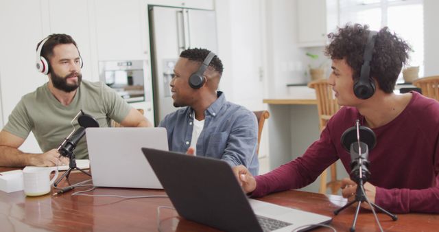 Three diverse men are seen podcasting at a table with laptops and microphones. They are engaged in conversation and wearing headphones. This stock photo is ideal for use in articles or websites related to podcasting, home office setups, audio production, teamwork, and digital media creation.