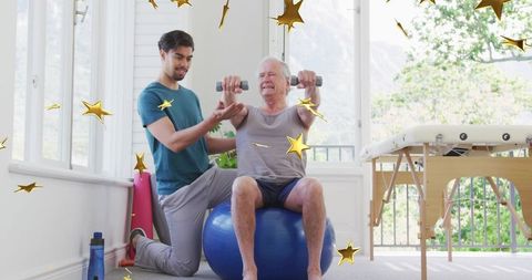 Senior man lifting dumbbells on stability ball while trainer assisting mobility and rehab