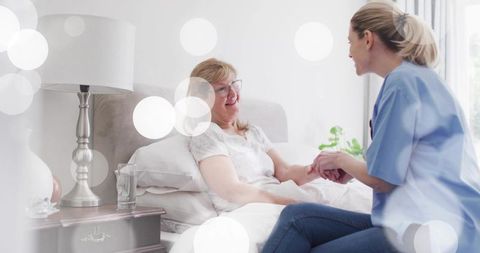 Caregiver in light-blue scrubs holding hands with smiling senior woman on bed at home
