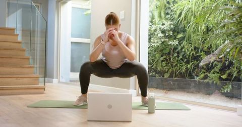 Woman doing deep squat during online workout at home on mat with laptop and water bottle