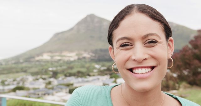 Young woman is smiling outdoors with a mountainous background. The image presents a joyful expression, natural setting, and relaxed atmosphere. Ideal for promoting travel, outdoor activities, well-being, and nature appreciation. Suitable for use in advertisement campaigns, blogs, social media posts, and lifestyle articles.