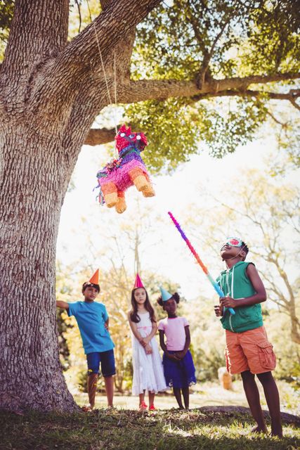 Group of children celebrating a birthday party in a park. One child, wearing a blindfold and holding a stick, is trying to break a colorful piñata hanging from a tree, while other children watch eagerly and wear party hats. Perfect for use in materials related to children's parties, celebrations, outdoor activities, and family fun.