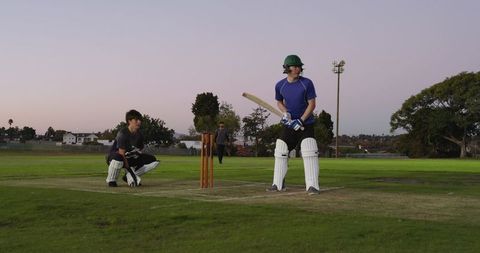 Teenage cricketer batting while bowler delivering and wicketkeeper crouching at dusk on suburban pit