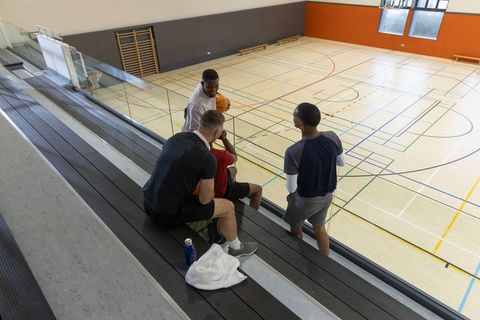 Diverse male teammates talking and sitting on gym bleachers overlooking basketball court
