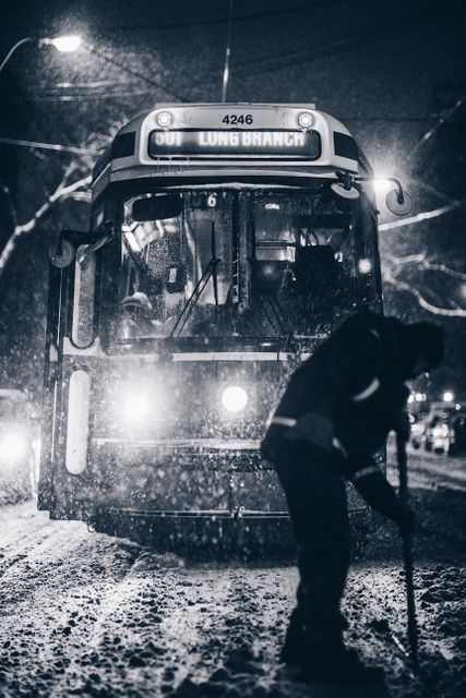 Night shot capturing a tram moving through a snowy urban street, with a worker seen shoveling snow. Ideal for use in themes of urban winter settings, public transportation, city life during snow, and road maintenance scenes. Suitable for illustrating cold weather conditions, snow removal efforts, and stories about urban transportation infrastructure.