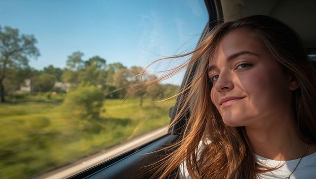 Sunlit young woman leaning out car window, wind-blown hair, countryside travel portrait