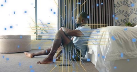 Contemplative man sitting on bedroom floor leaning against bed with digital data overlay