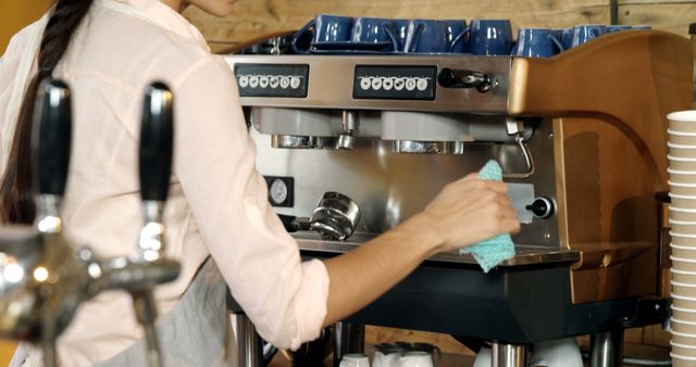 Barista wiping down an espresso machine with a cloth in a cozy coffee shop. Useful for illustrating themes related to coffee culture, maintenance of equipment, work environment, cleanliness, and café operations.