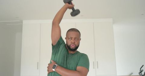 Man lifting dumbbell overhead while supporting arm wearing smartwatch during home workout