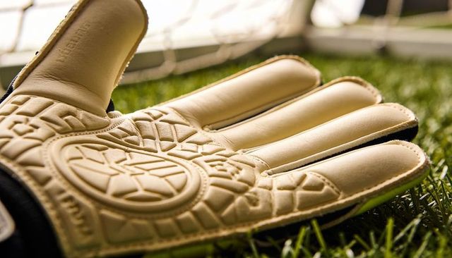 Close-up goalkeeper glove showing padded palm and grip on turf near goal net